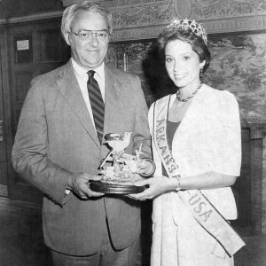 Older white man with glasses in suit and tie standing with young white woman in tiara and sash holding bird statue