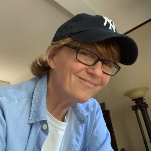 White woman with glasses smiling in New York Yankees hat and open denim button-up over white T-shirt