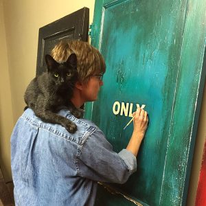 White woman with glasses in denim shirt painting doors while a clack cat rests on her shoulders