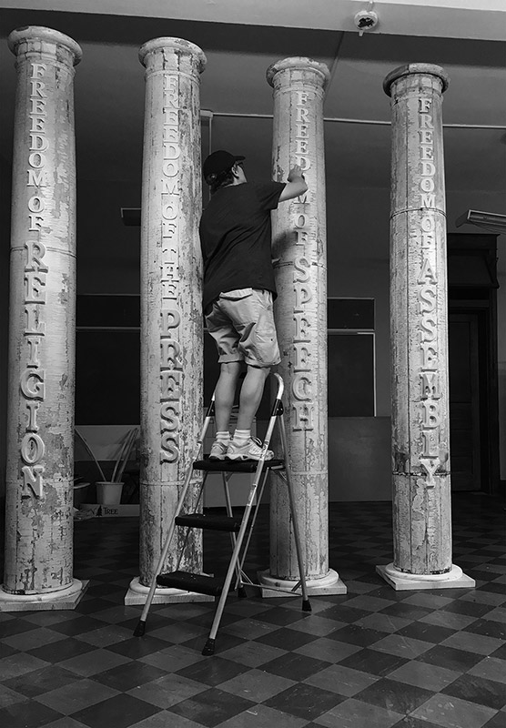 White woman in cap and shorts on ladder stool working on "Freedom of Speech" pillar in four pillar display