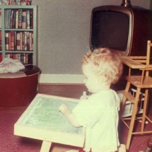 White baby girl playing with chalkboard in room with television high chair and bookshelf on red carpet