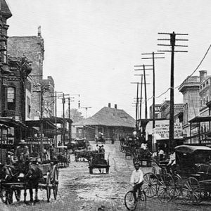 Horse drawn carriages on dirt road with multistory buildings on both sides