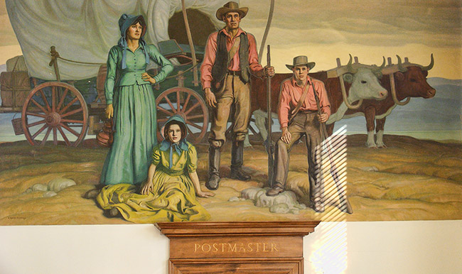 White men and women with covered wagon pulled by oxen in painting above "Postmaster" door