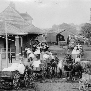 Crowd of white men with horse drawn wagons at single-story train depot building with outbuildings