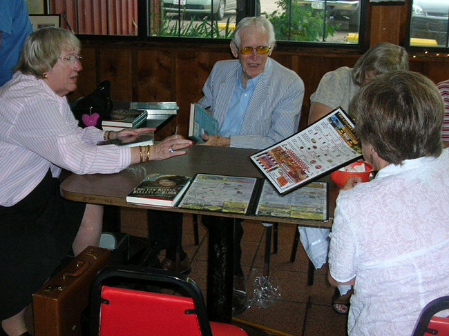 Older white man with sunglasses sitting at table with group of older women at book signing event