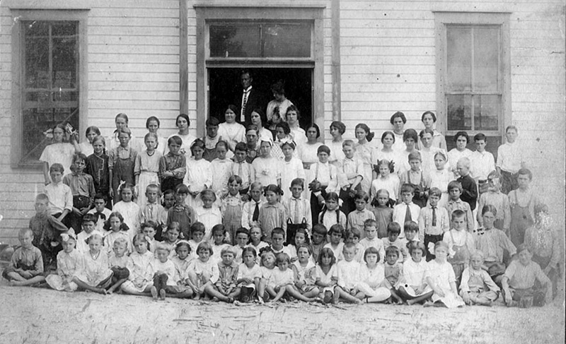 Large group of adults and children posing in front of wooden building