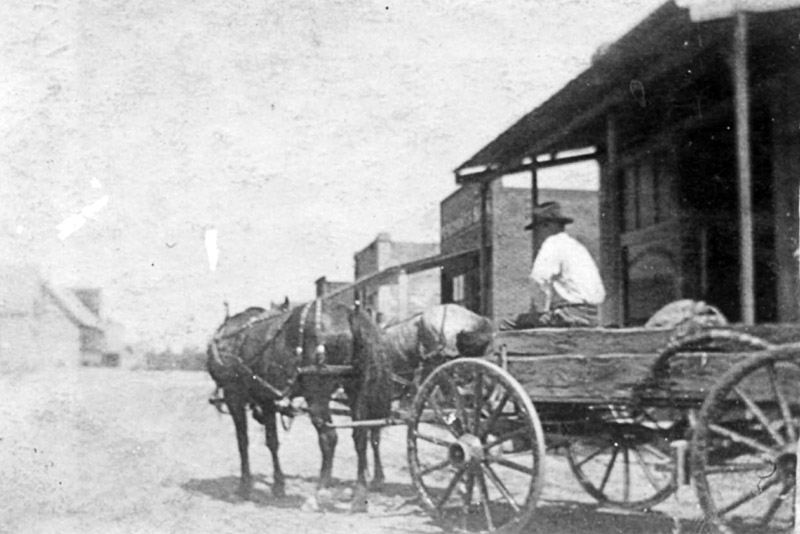 Man driving wagon and team of horses near wooden storefronts