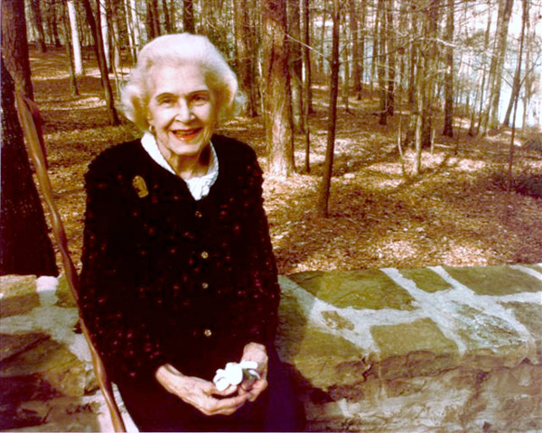 older white woman in cardigan on stone wall in front of woods