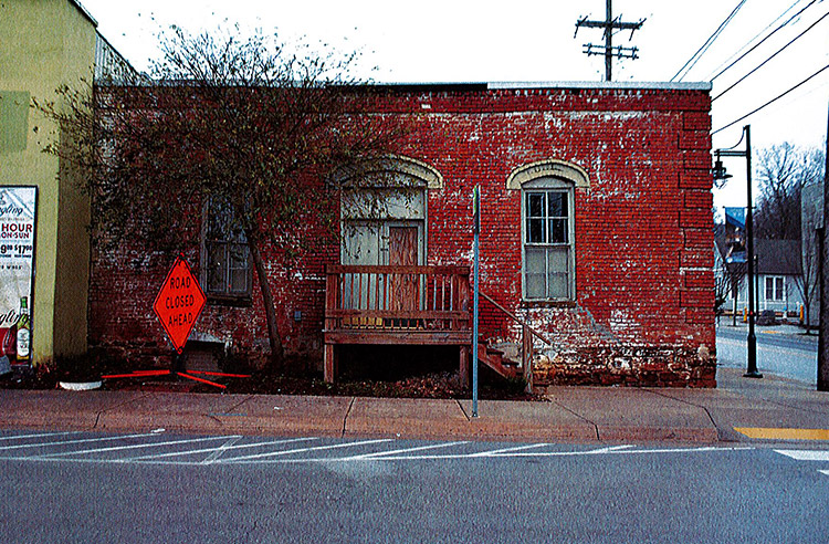 Single-story brick building with wooden stairs leading to entrance tree and "Road Closed" sign on sidewalk