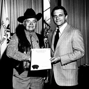 White man with cowboy hat in western clothing being handed a proclamation by white man in suit and tie