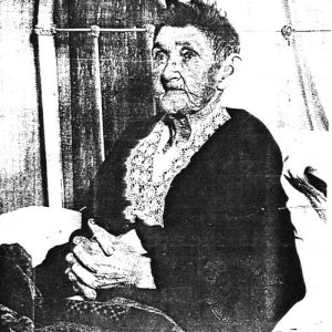 Older white woman sitting on bed in black dress with lace collar