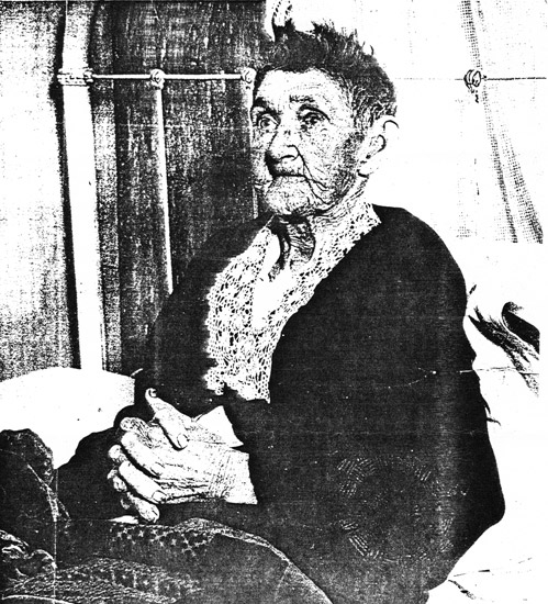 Older white woman sitting on bed in black dress with lace collar
