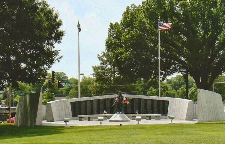 Memorial wall with plaque long stone bench and statue with flags and flowers