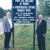 Older white man with beard and white man in suit and tie standing on either side of blue road sign with white text