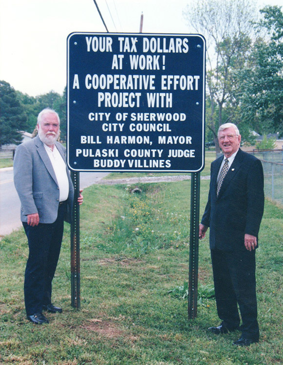 Older white man with beard and white man in suit and tie standing on either side of blue road sign with white text