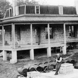 White man sitting in front of house with porch and barrels around him