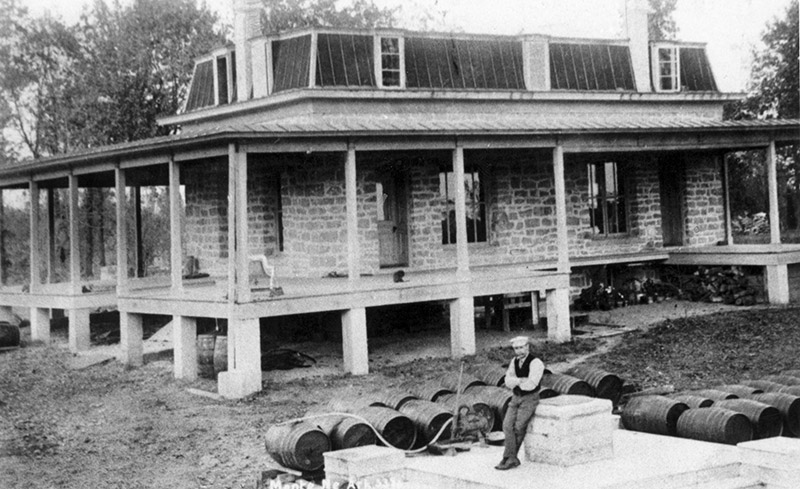 White man sitting in front of house with porch and barrels around him