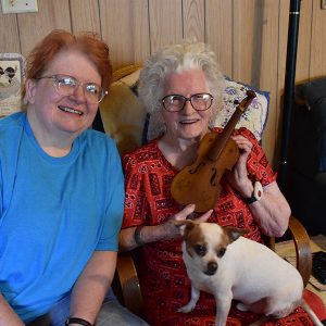 Old white woman with glasses holding a tiny fiddle next to white woman in glasses and blue shirt