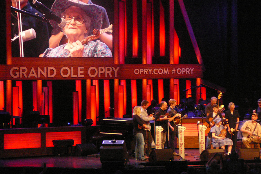 White musicians performing on stage with old white woman on screen behind them