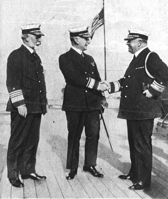 Three older white men in Naval uniforms with a flag behind them