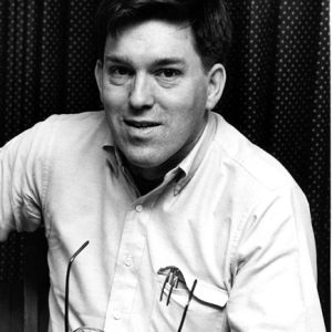 White man smiling in button-up shirt holding a pair of glasses in his right hand