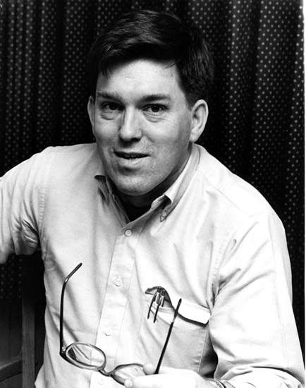 White man smiling in button-up shirt holding a pair of glasses in his right hand