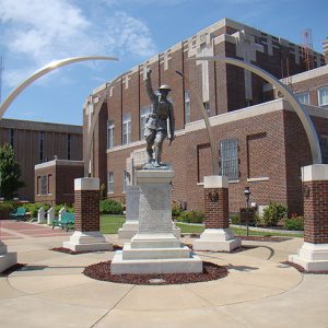 Statue of soldier on stone pedestal with brick columns with curved metal arms reaching toward center with a building in the background