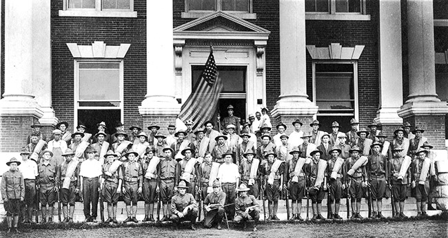 Group of white men in military uniforms with flag before brick courthouse with four columns