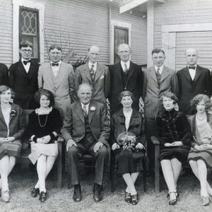 Posed group photo of white men in suits and women in dresses outside house