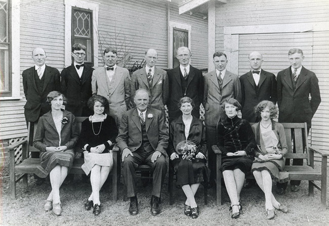 Posed group photo of white men in suits and women in dresses outside house