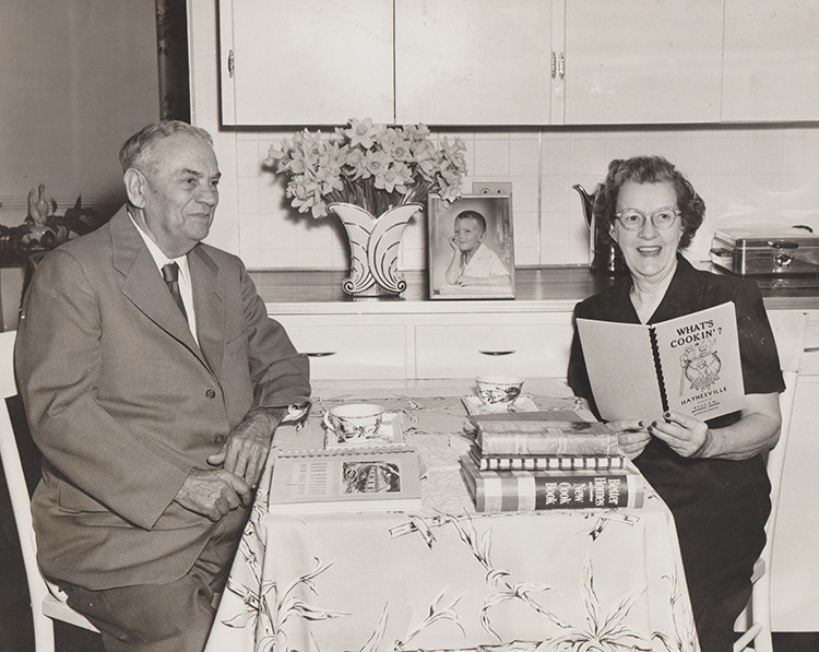 Old white man in suit and tie seated at kitchen table across from older white woman with glasses with cook book