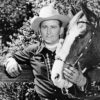 Portrait of a white man in western wear leaning on fence with his hand on a horse's head