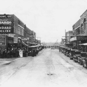 Line of parked cars in front of multistory brick storefronts on street with "Standard Gasoline" on the right side in the foreground