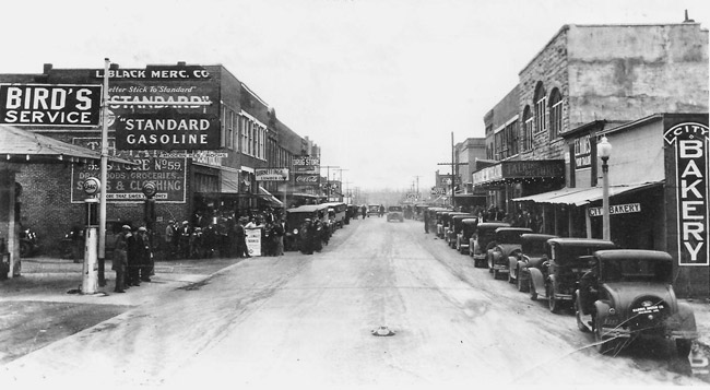Line of parked cars in front of multistory brick storefronts on street with "Standard Gasoline" on the right side in the foreground