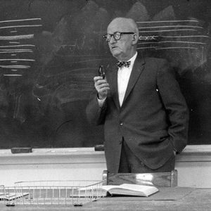 Older white man in suit speaking at chalkboard in classroom