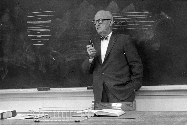 Older white man in suit speaking at chalkboard in classroom