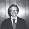 White man in suit and glasses standing in front of wood paneling