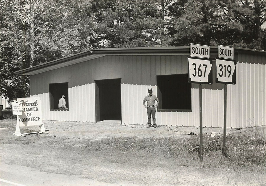 Single-story metal building with "Ward Chamber of Commerce" sign and white man in window and white man standing outside