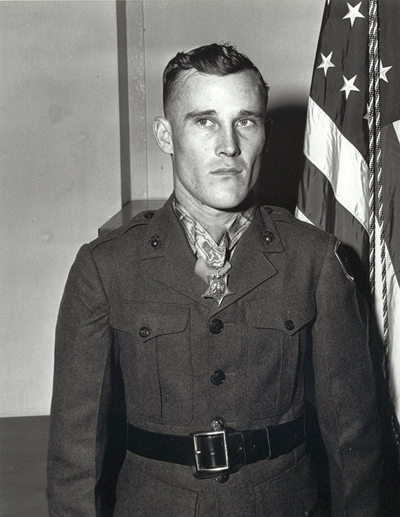 Young white man wearing Medal of Honor standing in military uniform with American flag behind him