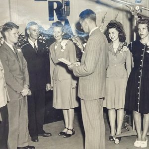 Group of young white men and women in studio with white man in suit standing before them