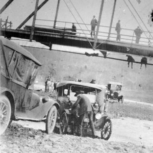 Men standing on and hanging from steel truss bridge with men with cars underneath it