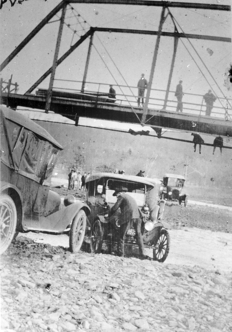 Men standing on and hanging from steel truss bridge with men with cars underneath it