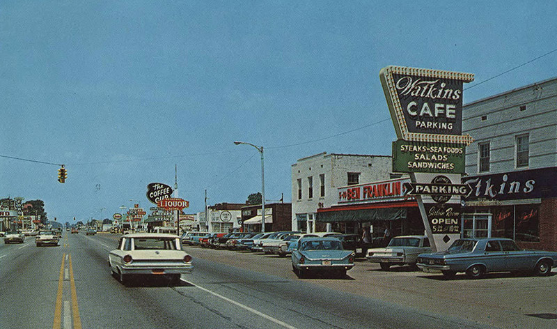 Traffic on two-lane street lined with stores and restaurants on both sides with signs and traffic lights