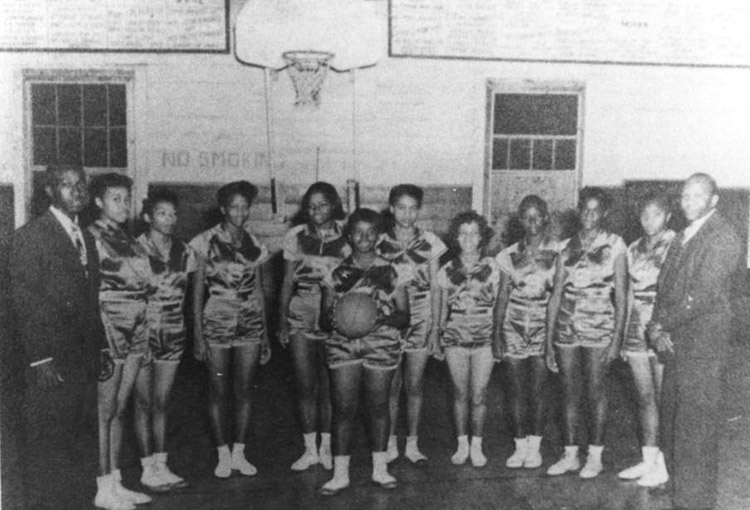 African-American girls in matching uniforms with African-American men in suits standing below basketball hoop