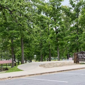 Covered benches and trees in park next to parking lot with "White Hall City Park" sign