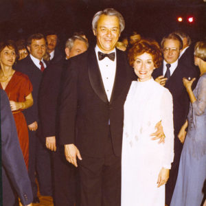 Older white man and suit and woman in white dress with white crowd behind them