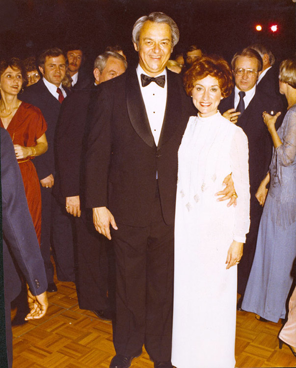 Older white man and suit and woman in white dress with white crowd behind them