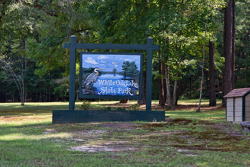 Bird and lake on "White Oak Lake State Park" sign in flower bed with trees behind it