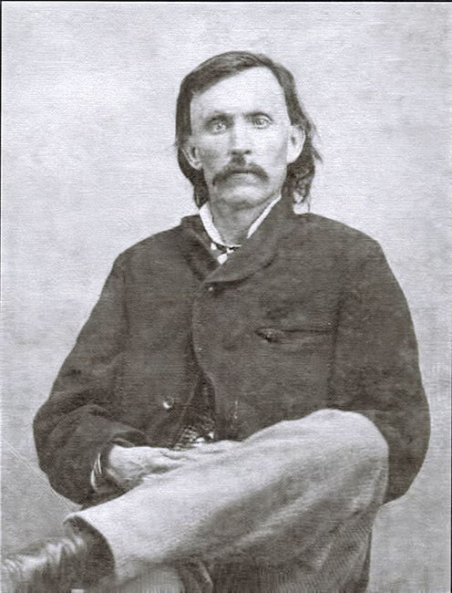 White man with mustache in sitting in suit with legs crossed