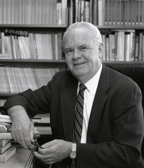 Older white man smiling in suit and tie sitting at desk with bookshelves behind him
